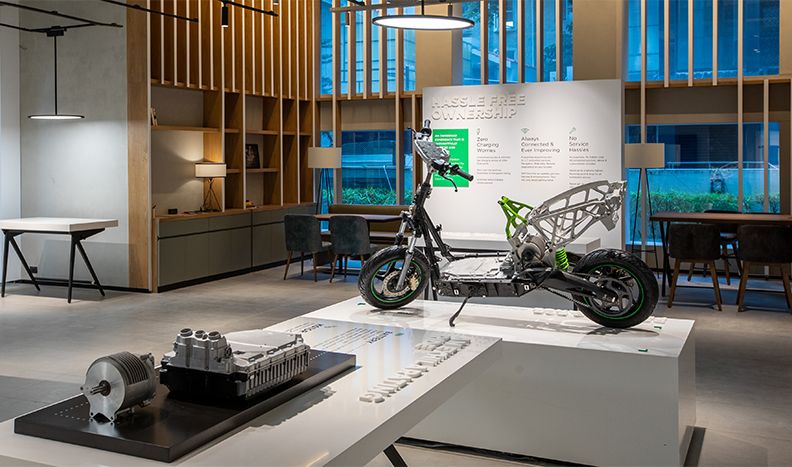 A stripped-down scooter is displayed on a platform inside the Ather showroom. A table in the foreground shows the motor assembly. 